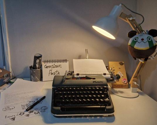 A photo of a desk. It has a typewriter on top of it, with a desk lamp shining light on the page. There are a few pages with text nearby, with some marker text over the text. The desk lamp has a plushie of an uglycute animal, Oopa mascot from a "Steins;Gate" game. A small notebook behind the typewriter says "Complain pasta" written in large cursive