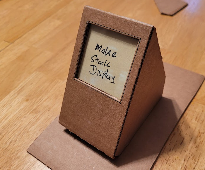 A cute little cardboard display for Post-It notes. It kind of looks like a little computer display. There is a clear plastic window in the cardboard face. It sits at an angle and displays exactly one mini Post-It note.
