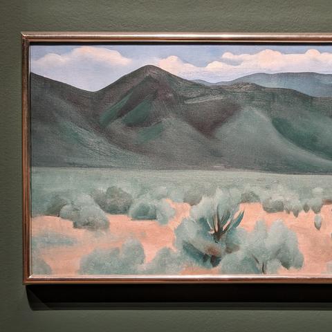 Image: A Georgia O'Keeffe painting hangs on the wall in the O'Keeffe and Moore exhibit at the Musee des Beaux Arts de Montreal, June 2024. The painting depicts a New Mexico landscape: foreground of high desert red soil, sage and mesquite receding an etched horizon of evergreen/black hills in the distance.