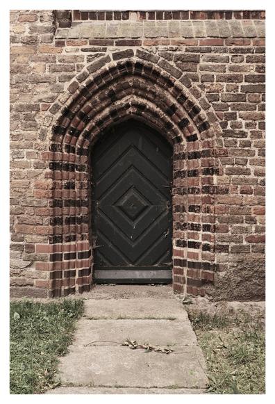 Eine schmale, spitzbogige Holztür mit diagonal angeordneten Brettern in einem gotischen Backsteinportal. Die Backsteine um die Tür sind in dekorativen Mustern angeordnet, teils dunkel glasiert. Vor der Tür führt ein schmaler, leicht verwitterter Steinweg durch einen Streifen Gras.