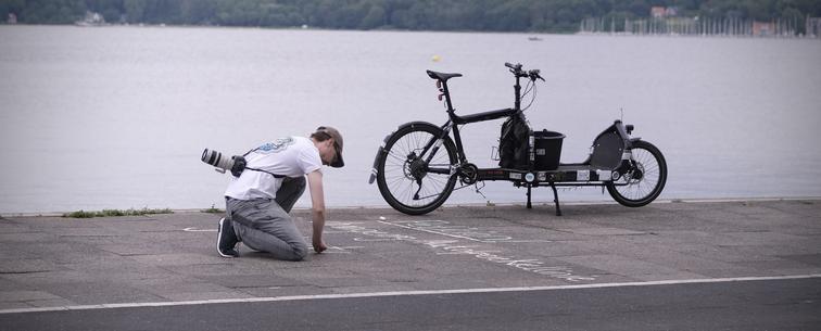 Eine Person kniet auf einem Weg am Wasser und schreibt mit Kreide auf den Gehweg. In der Nähe befindet sich ein Fahrrad mit Ladefläche. Der Hintergrund zeichnet sich durch ein ruhiges Gewässer und Grün in der Ferne am Kieler Ostufer aus.