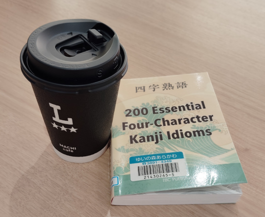 The small paperback library book has a Hokusai-esque wave painting on it and a library bar code. To the left is a black paper coffee cup with a black plastic lid. There is a large L above 3 stars on the cup. Both are on a table
