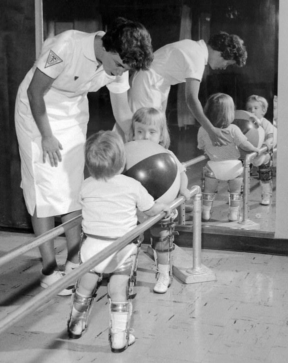This is a black and white photograph depicting a therapy session for children affected by polio. In the foreground, two young children are shown from behind, both wearing metal leg braces extending from their hips down to their feet. They are holding onto parallel bars, which are common equipment for gait training and physical therapy.  The child on the right is holding a large, two-toned (likely black and white or dark and light) inflatable ball in front of them, using it to lean against the parallel bars. Behind the two children, a woman in a white uniform, resembling a nurse or therapist, is bending over and assisting them. She has short, dark hair and is wearing a white dress or uniform with short sleeves, and a patch (possibly a Red Cross or medical symbol) on her right sleeve. Her expression is focused and caring as she looks at the children. To the right side of the image, a large mirror reflects the scene. In the reflection, we can see the front of the woman assisting, and the back of the child on the right, still holding the ball and wearing braces. Another child's reflection is also visible in the mirror, slightly behind and to the right of the first two, also wearing braces and looking towards the ball. The overall tone of the image is one of medical care, determination, and the challenges faced by children undergoing physical rehabilitation.