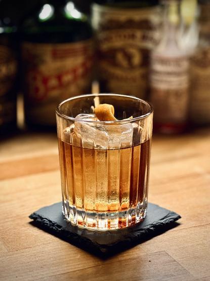 amber brown cocktail with a big rock of clear ice and orange twist garnish sitting on a black coaster with bottles in the background: Doctor Bird, Tempus Fugit Crème de Banana, Dutch Cacao Crème de Cacao, Peychaud’s Bitters, and Fee Brothers Black Walnut Bitters