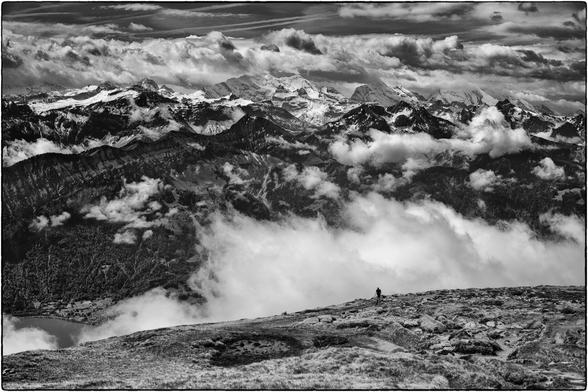 Schwarzweissfotografie der Berner Alpen mit Wolken und eines einzelnen Wanderers mit Stöcken.