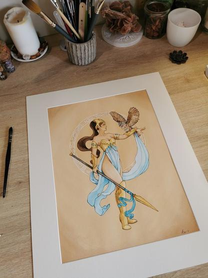 Sur un bureau, une aquarelle représentant Athéna avec une chouette sur fond ocre est sertie dans un passe-partout blanc cassé pour le faire ressortir une fois encadrée.