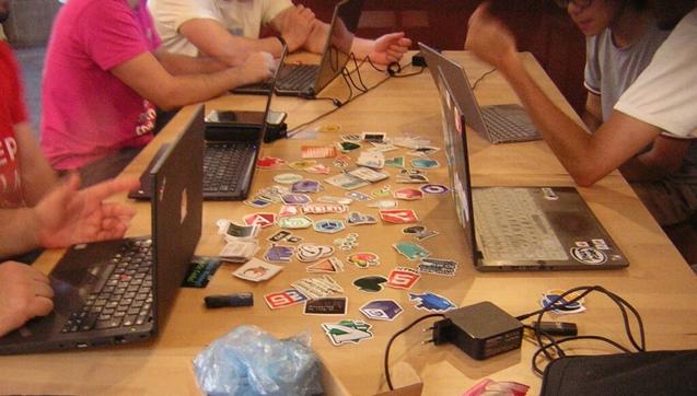 Un montón de pegatinas de software libre sobre una mesa. A los lados pueden verse varios portátiles y torsos de personas que parecen estar conversando en común.