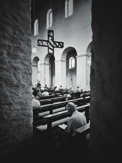 A black and white, vertical shot shows a church interior, looking down an aisle from between two stone pillars. In the foreground, the back of a man seated in a pew is visible as he reads. Other individuals are seated further down the pews, facing the front of the church. In the midground, a large, dark-toned acrylic cross hangs from the ceiling. Arched windows line the upper part of the far wall, and arched doorways are visible along the side.