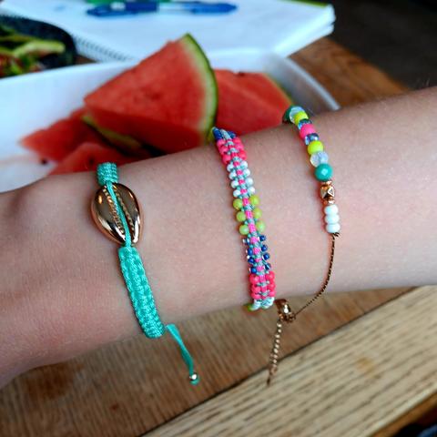 A small arm wearing three different summer bracelets of slightly different styles and colours. In the background you can see a wooden table and a bowl of watermelon.