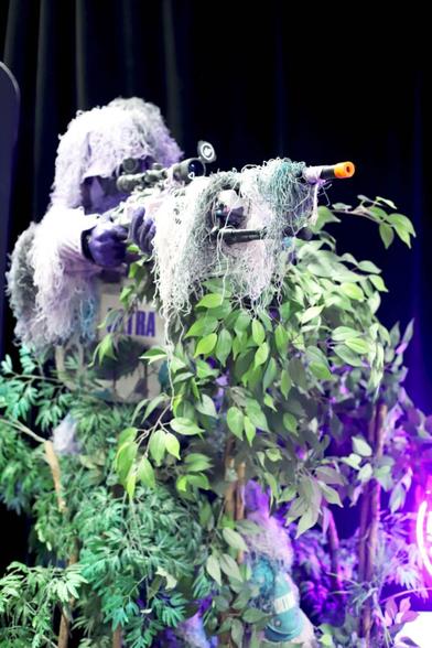 Photo of a life sized standing figurine dressed like a soldier in full jungle combat gear, including bundles of loose string for camouflage, posed behind some fake plants and pointing his toy weapon as if to shoot.