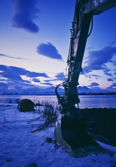 a backhoe rests in the snow on top of a hill overlooking the fjord and the mountains across the water, this is the 5 hours long blue hour period in winter beginning at about 10am and fades into pitch black 14 hour nights