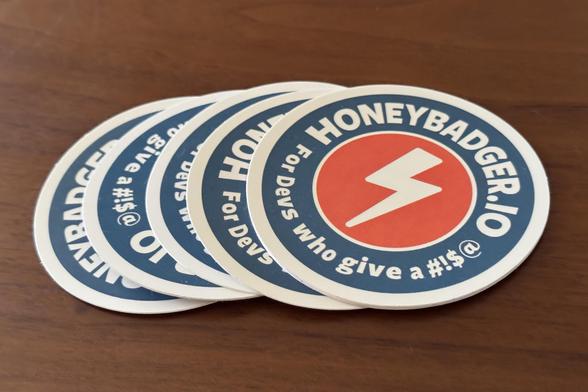 Stack of Honeybadger promotional stickers with the company’s orange and white lightning bolt logo and text reading ‘For Devs who give a #!$@’ on a blue circular background.
