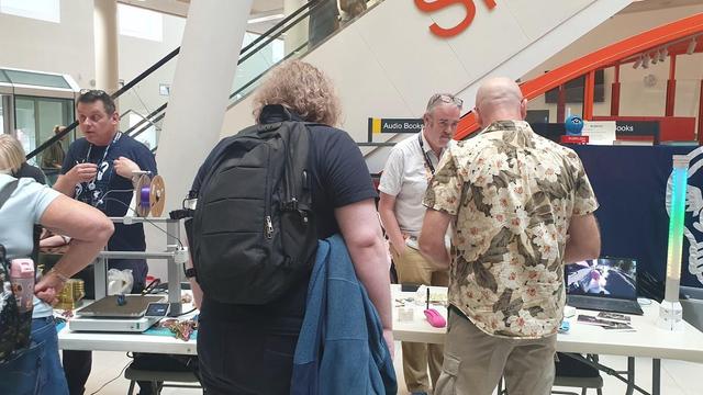 Adrian still stood behind that same table facing the camera, but this time with a visitor leaning over looking at 3D printed and postcard items on the table. Chris is also in shot at a table on Adrian's right, he is gesticulating to a visitor.
