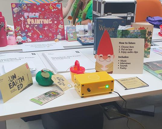 This is a photo of the Unearthed collection for Museum in a Box. It sists on a white table, with items such as a packet of seeds, a small fabric apple and a watering can. At the back of the table are accessibility guides for visitors. A display stand in the foreground advises people how to listen while in the background, a sign on another table says FACEPAINTING.