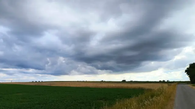 Eine ländliche Landschaft mit einem Feld voller grüner und gelber Pflanzen. Rechts verläuft eine schmale Straße entlang. Der Himmel ist bedeckt mit dunklen, bedrohlichen Wolken, die auf einen bevorstehenden Sturm hinweisen. In der Ferne stehen vereinzelt Bäume und ein einzelnes Windrad.