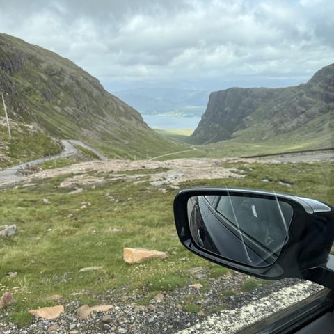 Photo taken from inside car with wing mirror visible, looking down the a winding road in the mountains with a sea loch in the distance.