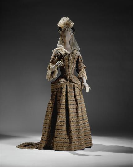 Two-piece wool dress, decorated with embroidery in silver-gilt thread. Lace sleeves extend from the elbows to the wrists, and there is an elaborate headpiece in lace and ribbon, with a lace trail to the shoulders.