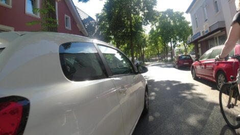 Ein weißer PKW überholt eine Radfahrerin mit zu wenig Abstand. Rechts parken PKW am Fahrbahnrand halb in Parkbuchten. Im schmalen Abstand zwischen der Radfahrerin und dem PKW ist offensichtlich noch das Fahrrad mit der mittig am Lenker montierten Kamera, welche das Bild aufgezeichnet hat.