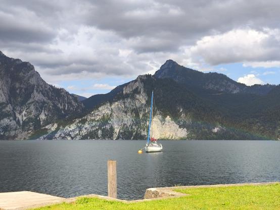 Ein Regenbogen ist über einem Boot am See zu sehen. Dahinter Berge. Im Vordergrund eine grüne Wiese.
