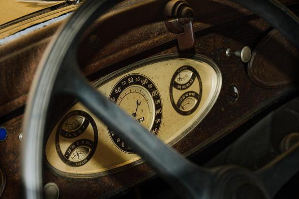 Vintage car dashboard with a large black steering wheel in the foreground. The dashboard features an oval speedometer displaying speeds from 0 to 90, flanked by two smaller round gauges. The left gauge reads "AMPERE" and the right gauge measures oil pressure in 100 to 600 kPa. The surface has a wood-like texture with signs of wear.
###
Oldtimer-Armaturenbrett mit einem großen schwarzen Lenkrad im Vordergrund. Das Armaturenbrett verfügt über einen ovalen Tachometer, der Geschwindigkeiten von 0 bis 90 anzeigt, flankiert von zwei kleineren runden Anzeigen. Das linke Manometer zeigt "AMPERE" an und das rechte Manometer misst den Öldruck in 100 bis 600 kPa. Die Oberfläche hat eine holzähnliche Textur mit Gebrauchsspuren.
