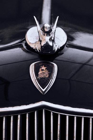 Close-up of a car hood featuring a chrome hood ornament shaped like a figure with wings, positioned above a shield-shaped badge. The badge displays text and an image of a ship, above a chrome grille with vertical bars. The car surface is glossy black.
###
Nahaufnahme einer Motorhaube mit einer verchromten Motorhaubenverzierung in Form einer Figur mit Flügeln, die über einem schildförmigen Abzeichen positioniert ist. Das Abzeichen zeigt den Text und das Bild eines Schiffes über einem verchromten Kühlergrill mit vertikalen Balken. Die Oberfläche des Autos ist glänzend schwarz.