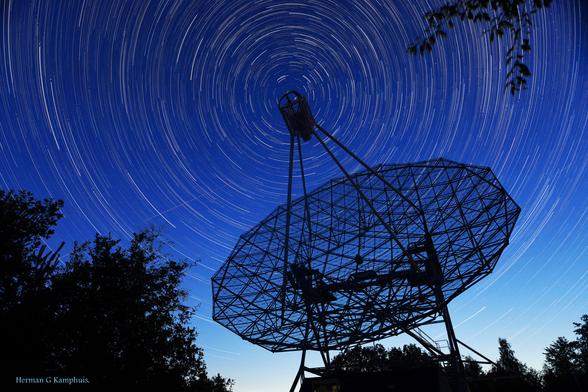 Nachtfoto van de radiotelescoop Dwingeloo. Door de lange belichting zijn de sterren uitgesmeerd in cirkels rondom de poolster ('startrails'). De poolster staat precies achter de focusbox van de telescoop.
