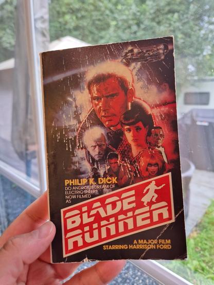 I am holding a book in front of a window. The cover says " Philip K. Dick Do Androids Dream of Electric Sheep, now filmed as Blade Runner, a major film starring Harrison Ford," with the film title more prominent and in the style from the movie poster.The book cover image is also from the poster of the film, featuring several people, spaceships and stylised rain drops.