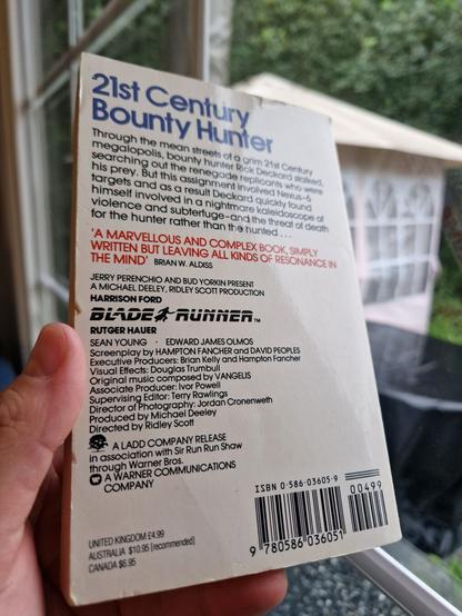 Holding the book again, now showing the back cover:

"21st Century Bounty Hunter 

Through the mean streets of a grim 21st Century megalopolis, bounty hunter Rick Deckard stalked, searching out the renegade replicants who were his prey. But this assignment involved Nexus-6 targets and as a result Deckard quickly found himself involved in a nightmare kaleidoscope of violence and subterfuge - and the threat of death for the hunter rather than the hunted... 

A marvellous and complex book, simply written but leaving all kinds of resonance in the mind - Brian W. Aldiss"

Then some details about the movie cast and crew that won't fit in the alt text, but you could look it up if you're curious.

Then original recommended sales prices:

United Kingdom: £4.99
Australia: $10.95 (recommended)
Canada: $6.95

Followed by the ISBN and barcode.