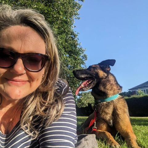 Selfie of a woman lying on grass in bright sunshine. Over her shoulder can be seen a dog who's looking away from the camera. Behind them both can be seen some trees and bright blue sky.