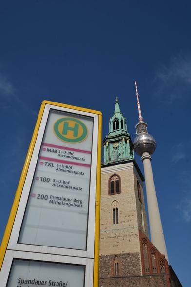 The image captures a city scene featuring a public transportation sign in the foreground, displaying routes and stops. Behind it, a historic church with a green spire and the iconic Fernsehturm stand tall against a clear blue sky.