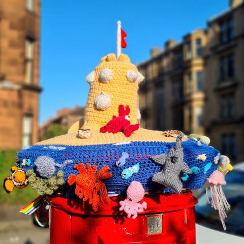A seaside-themed knitted postbox topper featuring a sandcastle in the middle and a variety of sea life around the sides.