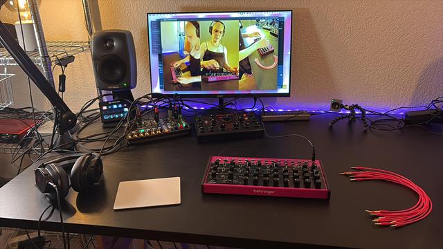 Behringer Edge, a pink synth on a black table. In the background, a computer screen is visible with three portrait oriented camera shots showing the girl operating the synth a bit earlier. Also in the frame, headphones and a portable mixer and some studio monitors, as well as purple LED strip lighting.