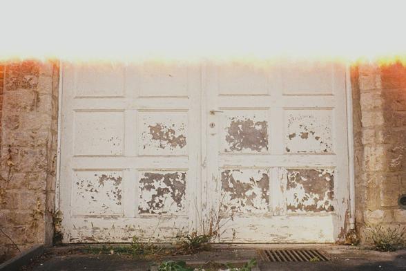 This image captures a weathered and aged garage door, composed of white-painted wooden panels, many of which are peeling and chipped with time. The door is set into a stone and brick wall, suggesting an older structure. Signs of neglect are visible in the overgrown weeds at the base and the small patch of gravel garden in front of the entrance. A light leak effect at the top of the photo washes out the upper portion of the image with a warm, glowing flare, adding a nostalgic and accidental aesthetic touch. The overall atmosphere evokes quiet decay and forgotten places, making it visually compelling in its stillness and imperfection.