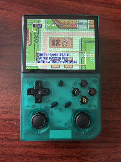 A translucent teal-green GameBoy-looking thing sits on a wood-grain tabletop. The device has SNES button layout and analog sticks below that. On the screen is Legend of Zelda: A Link to the Past and Link has just purchased the bottle from the merchant in the village.