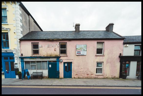 Street view of colourful terraced buildings in an Irish town, featuring a pink two-storey building with "McCARTHY'S" painted on its facade and a "SOLD" sign from "DNG" estate agents, flanked by a blue "SHELL SERVICE" shop on the left and other commercial premises.
