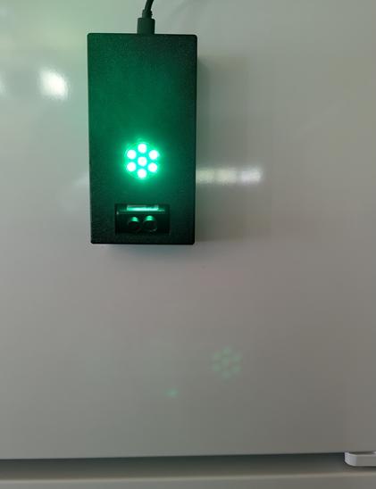A black plastic case is attached to the front of a white refrigerator. In the center of the front of the case is an LED ring consisting of 7 LEDs with one in the center surrounded by 6 in a ring around it. The LEDs are glowing green. Near the bottom of the case, a sensor protrudes slightly from the front. It resembles a tiny pair of binoculars, two lenses in a horizontal arrangement.