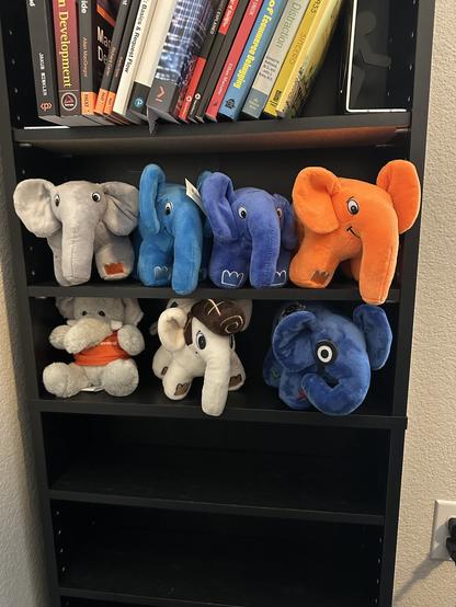 A bookcase with books on one shelf and multiple elePHPant plushies on the two shelves below it.