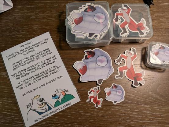 Photo of many sticker prints using the custom artwork from this post (orange fox curled up under weighted blanket and fox sprawled out on back blushing and tail wagging).

To the left lays a thank-you note from Bear and Beagle store for having ordered, offering support, etc.