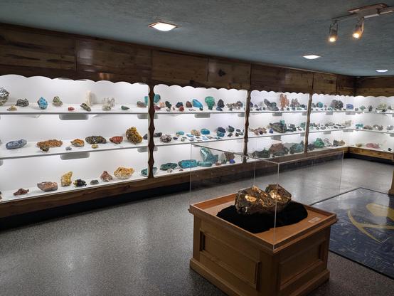 Lit up display case of a wall of various gem and mineral samples aligned on a wall.  Lots of bright blues, greens, red, as well as more traditional rock colors.  In the foreground is a massive piece of pyrite.