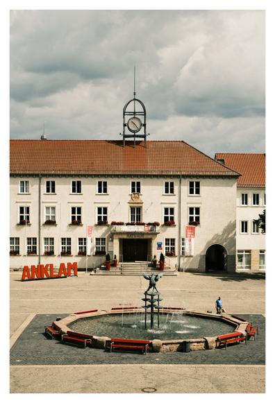 Auf diesem Foto sieht man den Marktplatz von Anklam, einer Stadt in Mecklenburg-Vorpommern, Deutschland. Im Vordergrund ist ein achteckiger Brunnen mit einer Bronzefigur in der Mitte zu sehen, umgeben von roten Bänken. Dahinter steht das weiße Rathaus mit roten Dachziegeln, Fensterkästen voller Blumen und einem modernen Uhrturm auf dem Dach. Links vor dem Rathaus steht eine große rote Buchstabeninstallation mit dem Stadtnamen „ANKLAM“. Über dem Eingang hängt ein Banner, das auf eine Jubiläumsfeier hinweist.