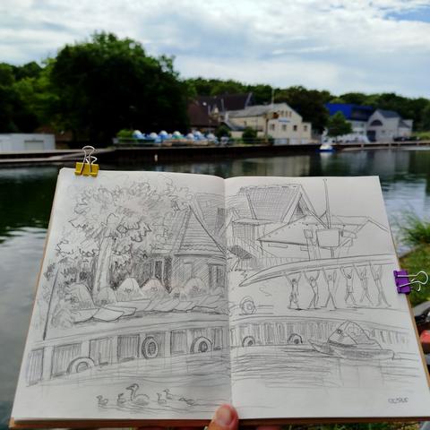 Skizzenbuch vor dem Kanal, im Hintergrund Boote, Häuser, Bäume.