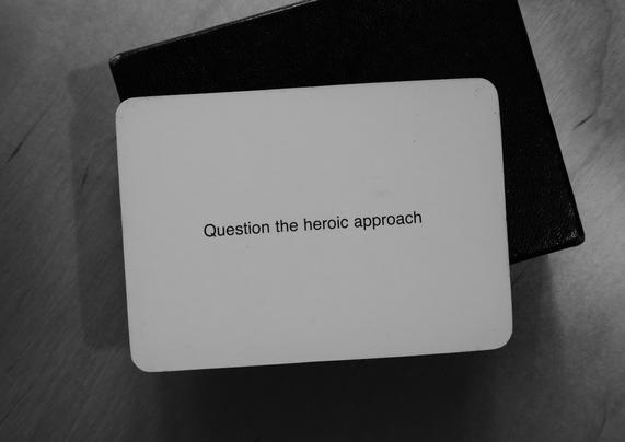 A black-and-white photo of an Oblique Strategy card. The card reads: 'Question the heroic approach'.