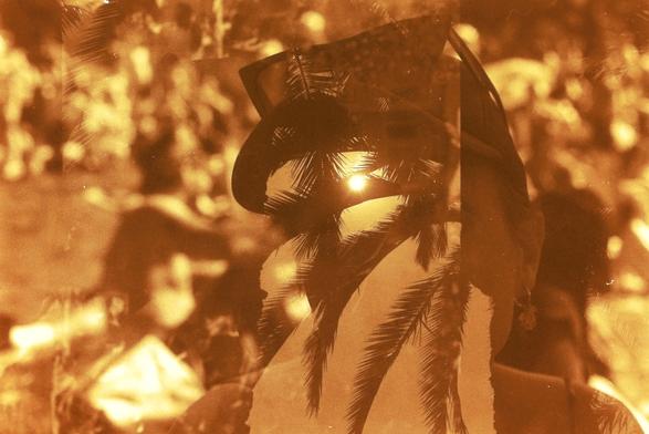A sepia-toned image with a beautiful woman in the foreground. She’s wearing an earring, a baseball cap, and has angular sunglasses perched on her head. In the background, blurred figures also enjoying the park on a sunny afternoon. In the center of the image a double-exposure of the sun and a palm tree obscures our view of that beautiful woman