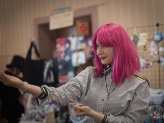 In the image, a young woman with pink hair is captured in mid-motion. She is wearing a striped gray shirt and has her arms outstretched, as if she's about to make an offer or gesture towards something. Behind her there is a little store in a form of a table with a merchandize placed on it. The store appears to be selling anime-related items.

The setting appears to be indoors. The overall atmosphere suggests a some sort of event with a particular interest in Japanese pop culture.