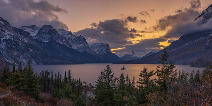 A sunset scene of a lake encircled by snow drizzled mountain peaks on the sides and trees in the foreground. The sky is shades of orange and the clouds are backlit fluffy cotton candy. The middle of the lake has a small island with a handful of trees.