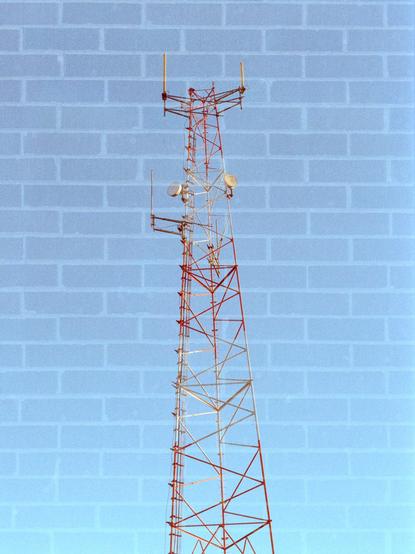 In camera double exposure: A radio tower made out of lattice construction and painted red and white with several dish and cylindrical antennas sticking out against a blue sky is super imposed on top of a brick background.
