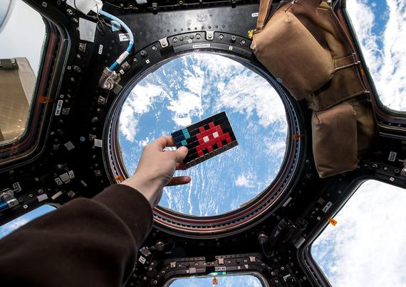 Mosaico de space invaders con un marcianito rojo frente a la ventana circular de la estación espacial internacional.
