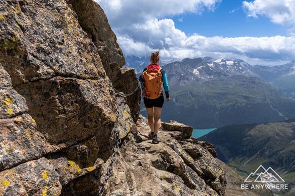 Wanderin auf schmalem Felsweg mit Kette am Piz Julier, mit Blick auf tiefblauen See, grüne Täler und schneebedeckte Berggipfel im Engadin.
