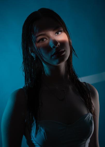 A young woman with wet hair looks slightly to the side, wearing a white strapless top. Her face is partially illuminated with a soft shadow across her eyes, while the background features a gradient of deep teal and dark blue. A faint diagonal light band is visible in the background.