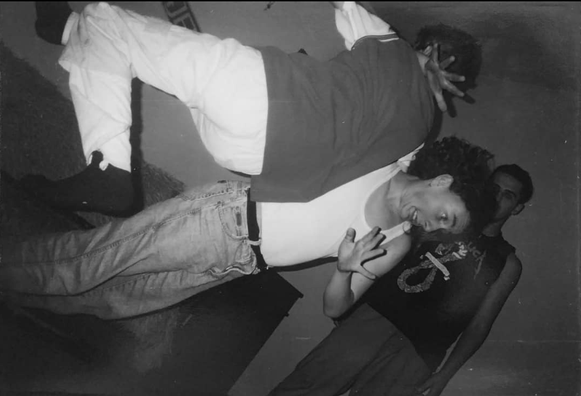 The image is a black and white photograph featuring three individuals in a dynamic pose. The central figure is leaning back with their head tilted to the side, wearing a white sleeveless shirt and light blue jeans. They are making a hand gesture near their face. The person behind them is wearing a dark shirt with a graphic design and dark pants, with their hand raised. The third person is partially visible, wearing a dark shirt and pants, standing behind the central figure. The background is plain and dark, with no distinct features. The lighting is low, creating shadows and emphasizing the figures' movements.

Provided by @altbot, generated privately and locally using Ovis2-8B